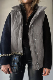 Quilted stand-collar utility gilet in Dust Storm with zip and snap closure, worn over a black sweater.