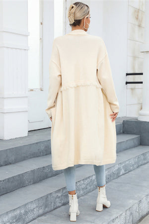Ivory longline knit cardigan with fringe trim and pockets, styled with light blue jeans and white boots.