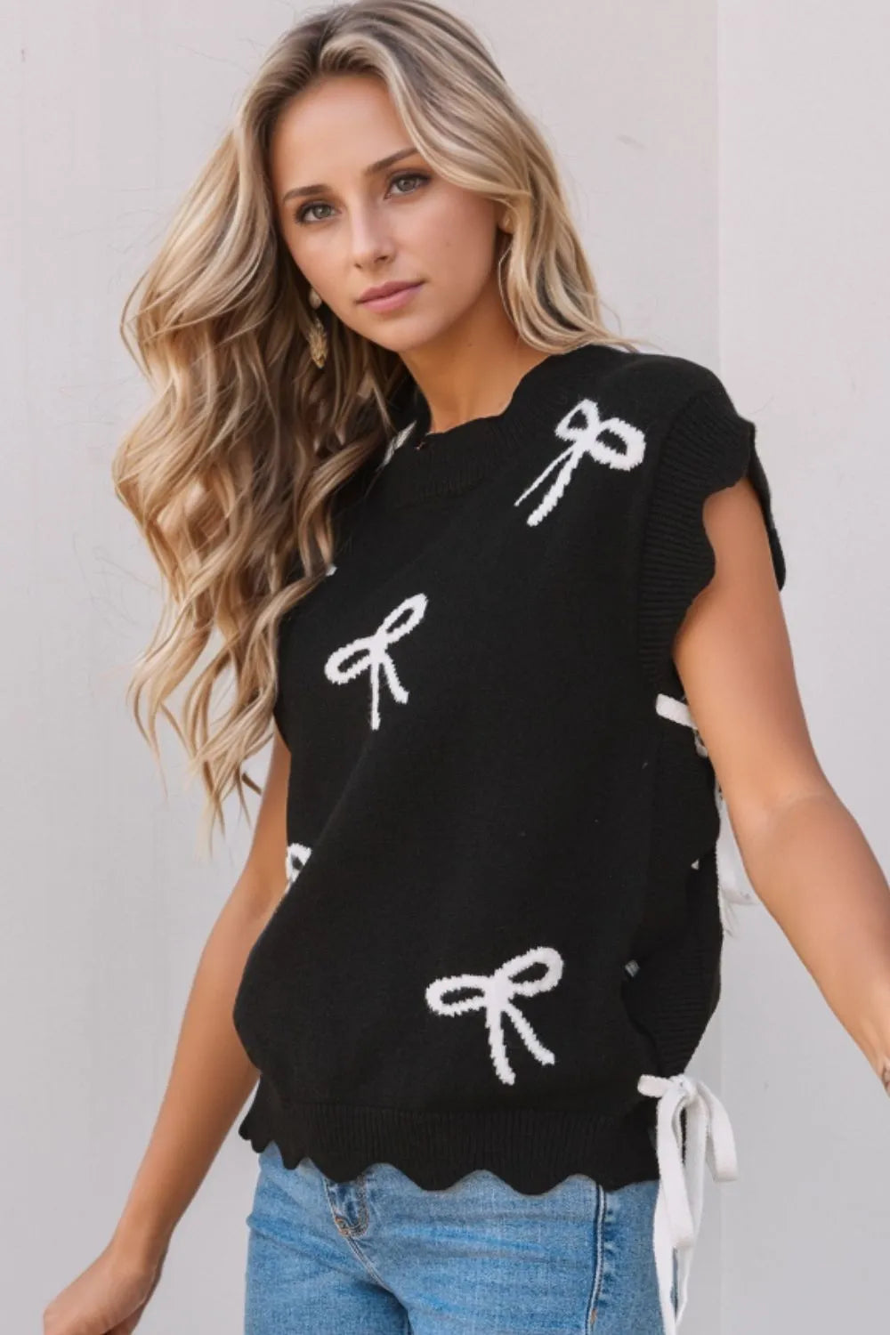 Black knit vest with white bow print, scalloped hem, and tie details.