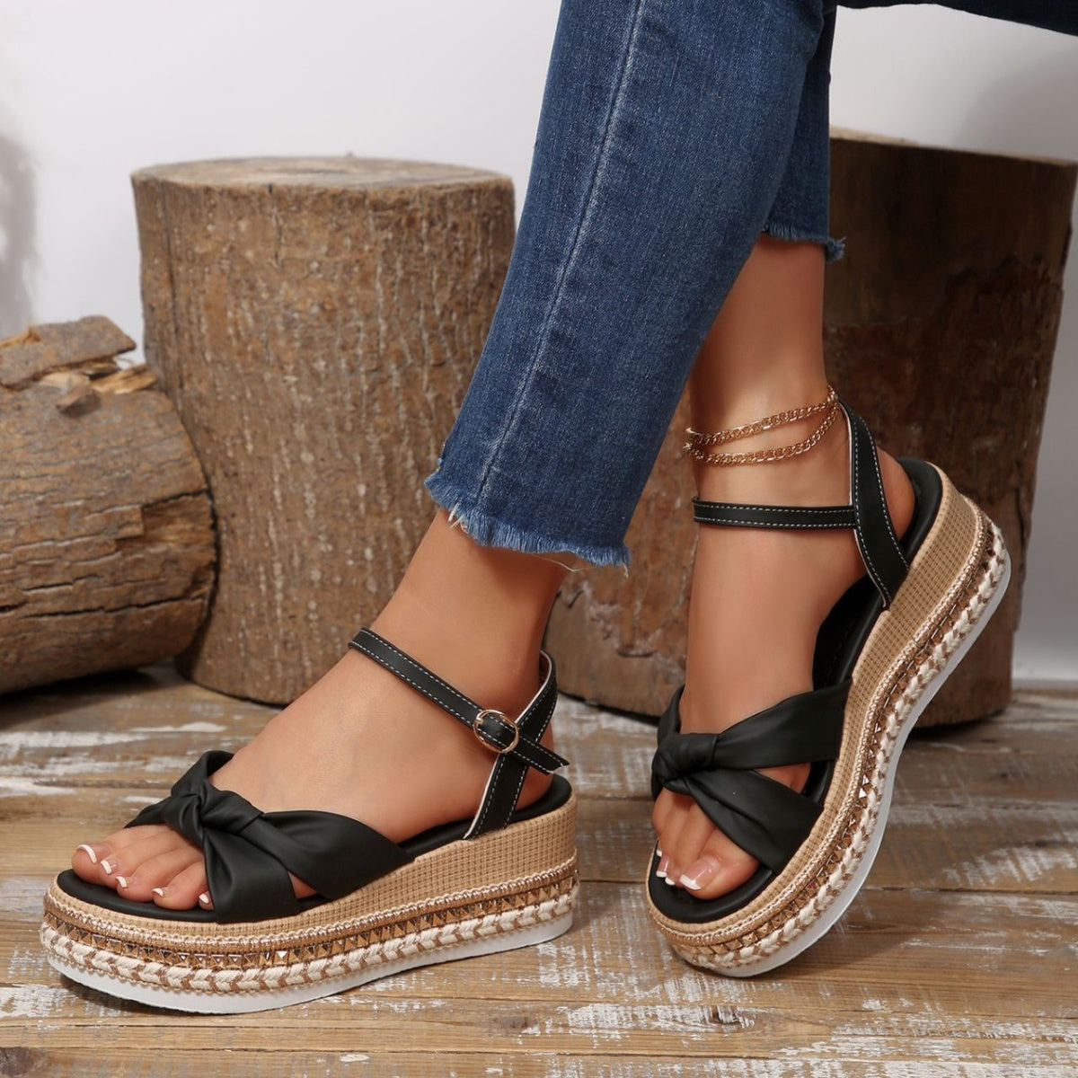 Black wedge sandals with bow knot detail and braided trim, featuring ankle buckle and gold chain anklet.