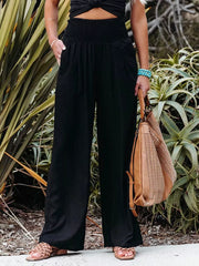 Black smocked wide-leg maternity pants with pockets, styled with sandals and a woven bag.