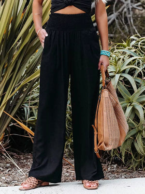 Black smocked wide-leg maternity pants with pockets, styled with sandals and a woven bag.