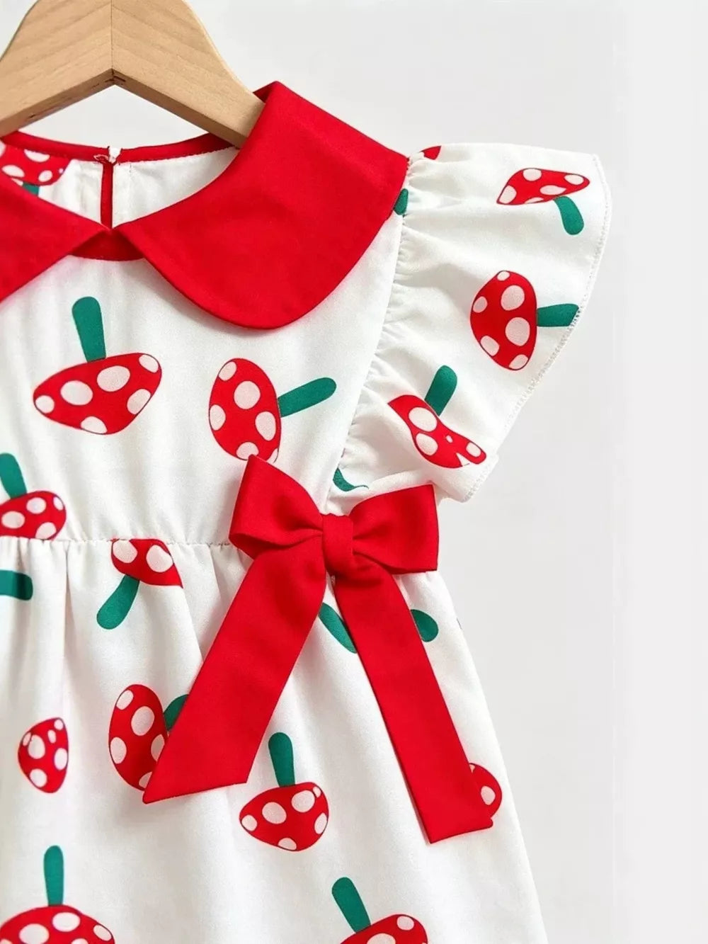 Baby set with white fabric, red mushroom print, Peter Pan collar, flutter sleeves, and red bow detail.