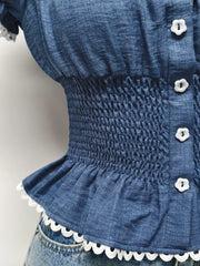 Blue denim maternity blouse with smocked waist, puff sleeves, decorative buttons, and ruffled hem.