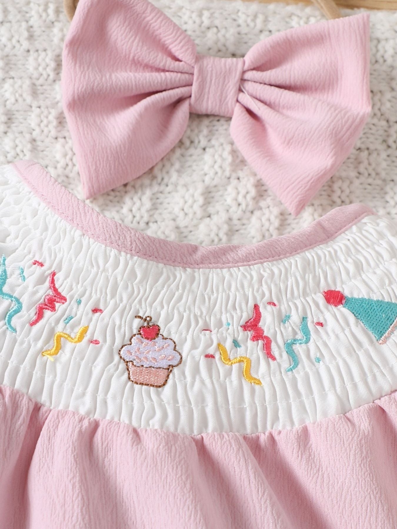 Blush pink baby one-piece with smocked white bodice featuring embroidered cupcake and confetti.