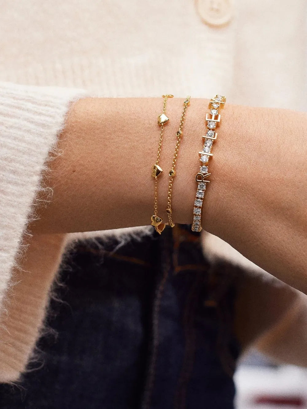 Gold chain bracelets with heart charms and a diamond tennis bracelet with initial charms.