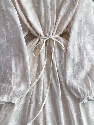 Elegant tie-waist dress in tan with sheer floral embroidery and long sleeves.