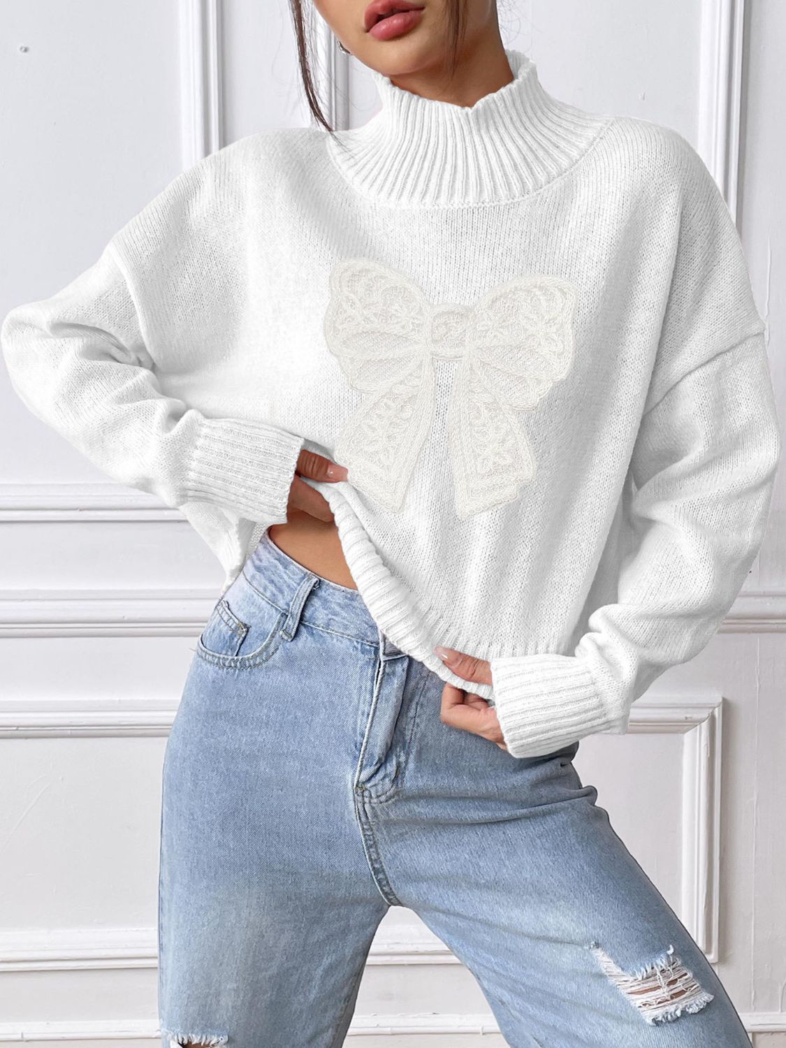 White turtleneck knit sweater with lace bow appliqué and ribbed cuffs.