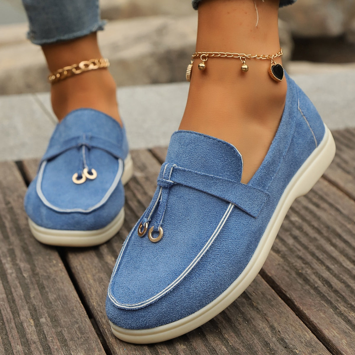 Blue suede flat slip-ons with decorative strap and tassel detail.