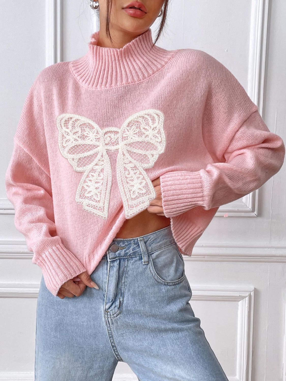 Blush pink turtleneck knit sweater with white lace bow appliqué.