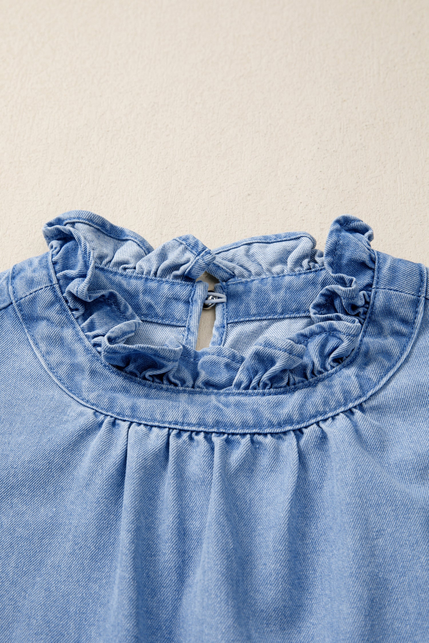 Light wash denim blouse with ruffled collar and button closure at back neck.