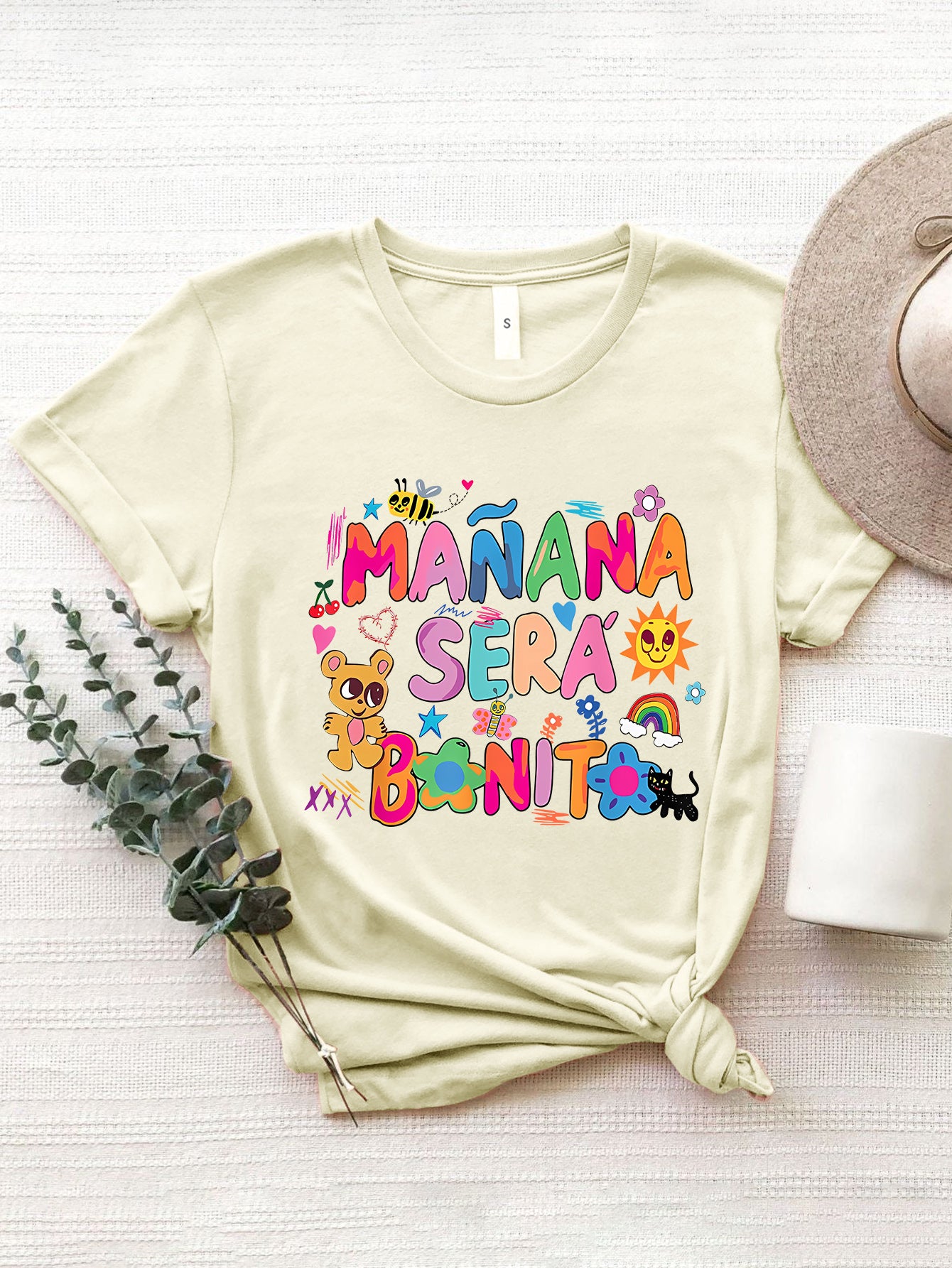 Cream graphic tee with "Mañana Sera Bonito" text and colorful cartoon illustrations.