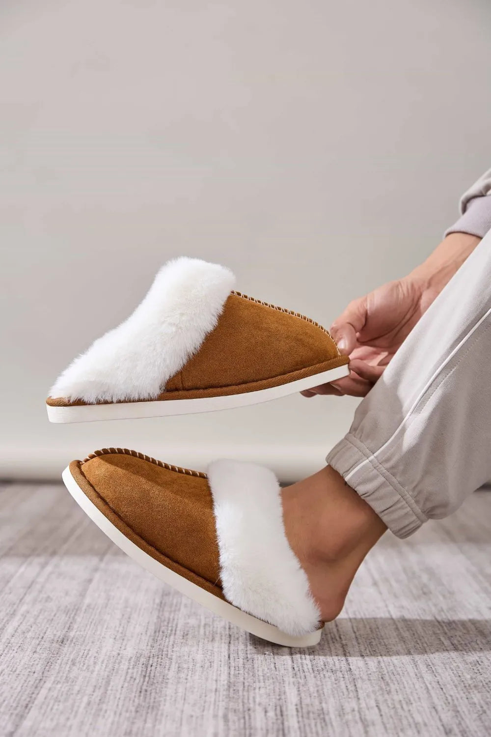 Caramel suede house slides with white shearling trim, one on foot, one held.