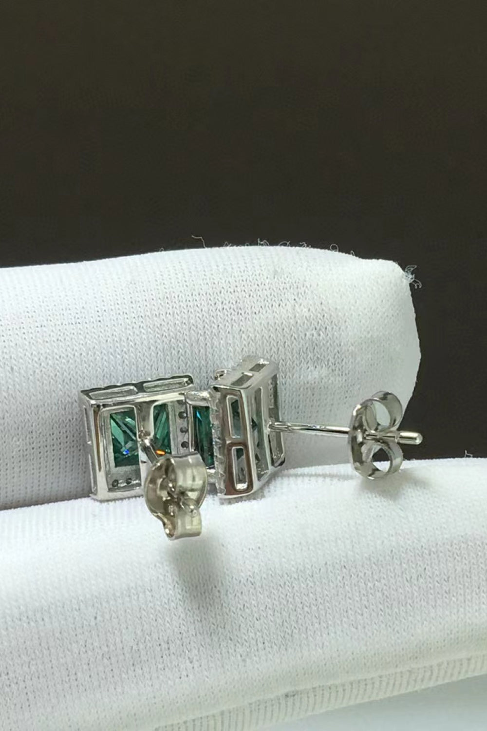 Green emerald cut stud earrings with diamond accents on a white cloth.