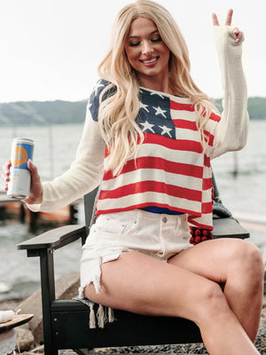 Ivory off-shoulder knit sweater with American flag print, long sleeves, and a relaxed fit.
