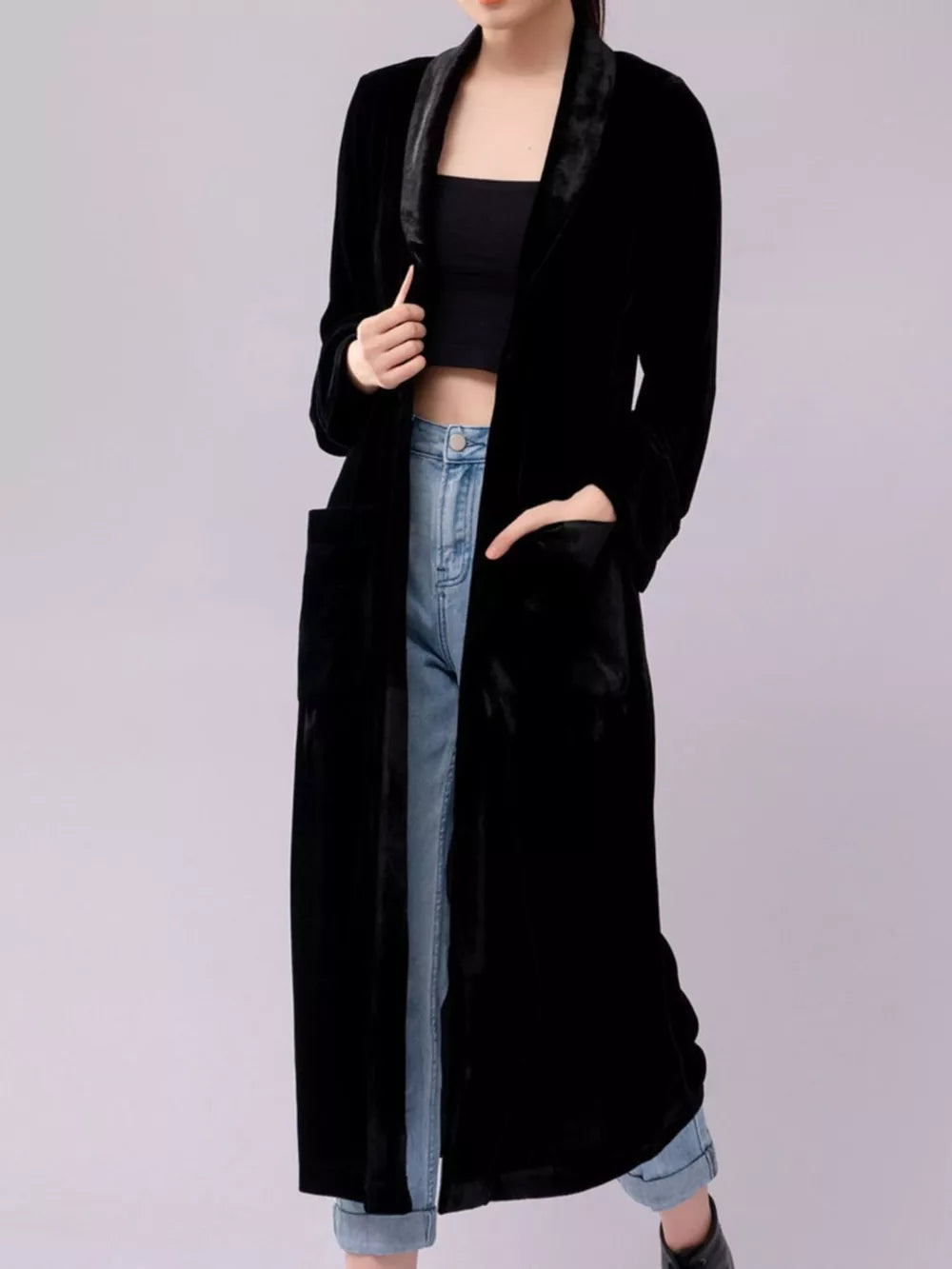 Black velvet longline trench coat with draped lapels and pockets, worn open over a crop top and jeans.