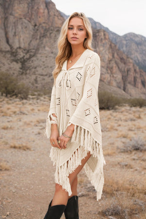 Cream knit poncho with geometric cutouts and fringe detail, featuring a tie neckline.