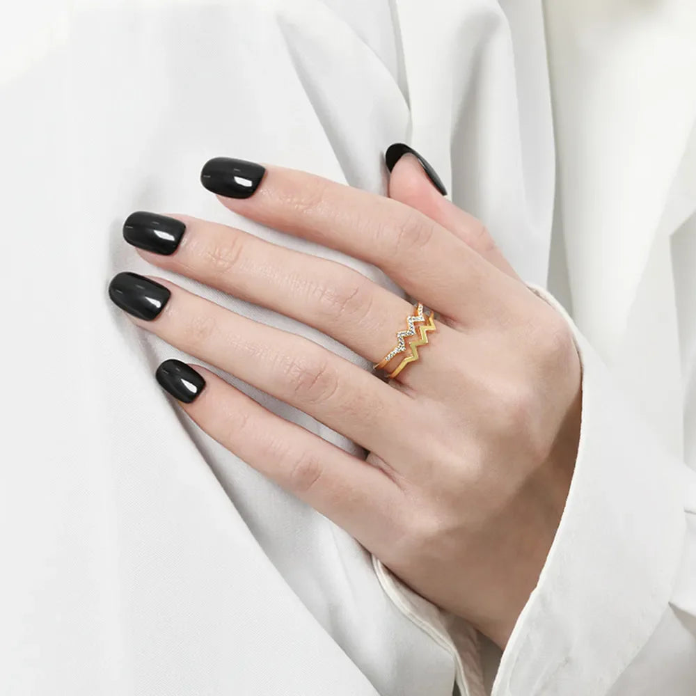 Gold zigzag split band ring with small clear stones, worn on a hand with black nails.