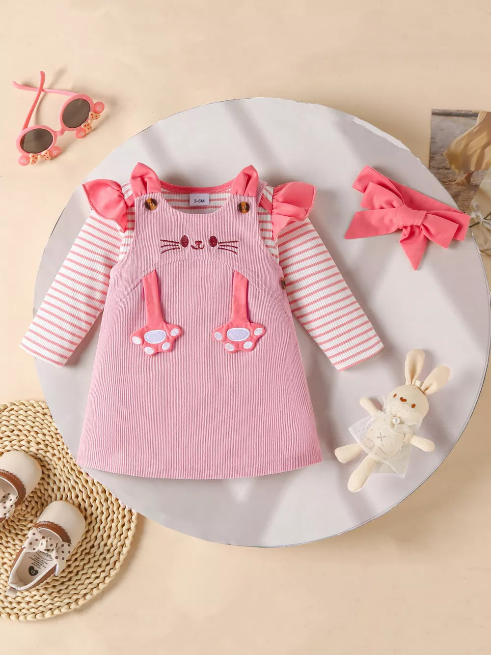 Pink corduroy cat applique pinafore dress with striped long sleeve bodysuit and ruffled shoulders.