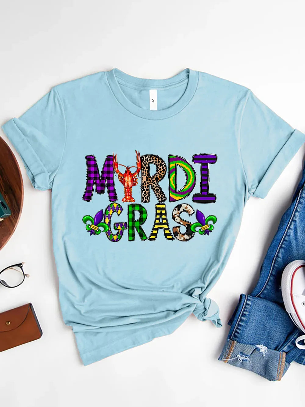 Light blue graphic tee with Mardi Gras text in mixed patterns and a crawfish graphic.