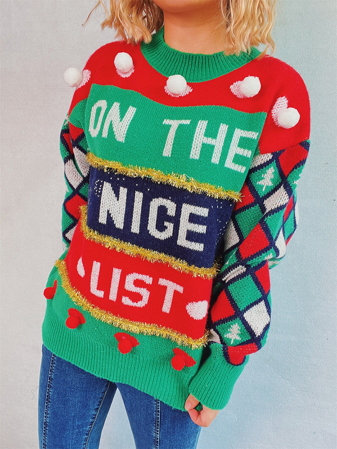 Green acrylic crewneck sweater with "On The Nice List" graphic and pom-pom details.