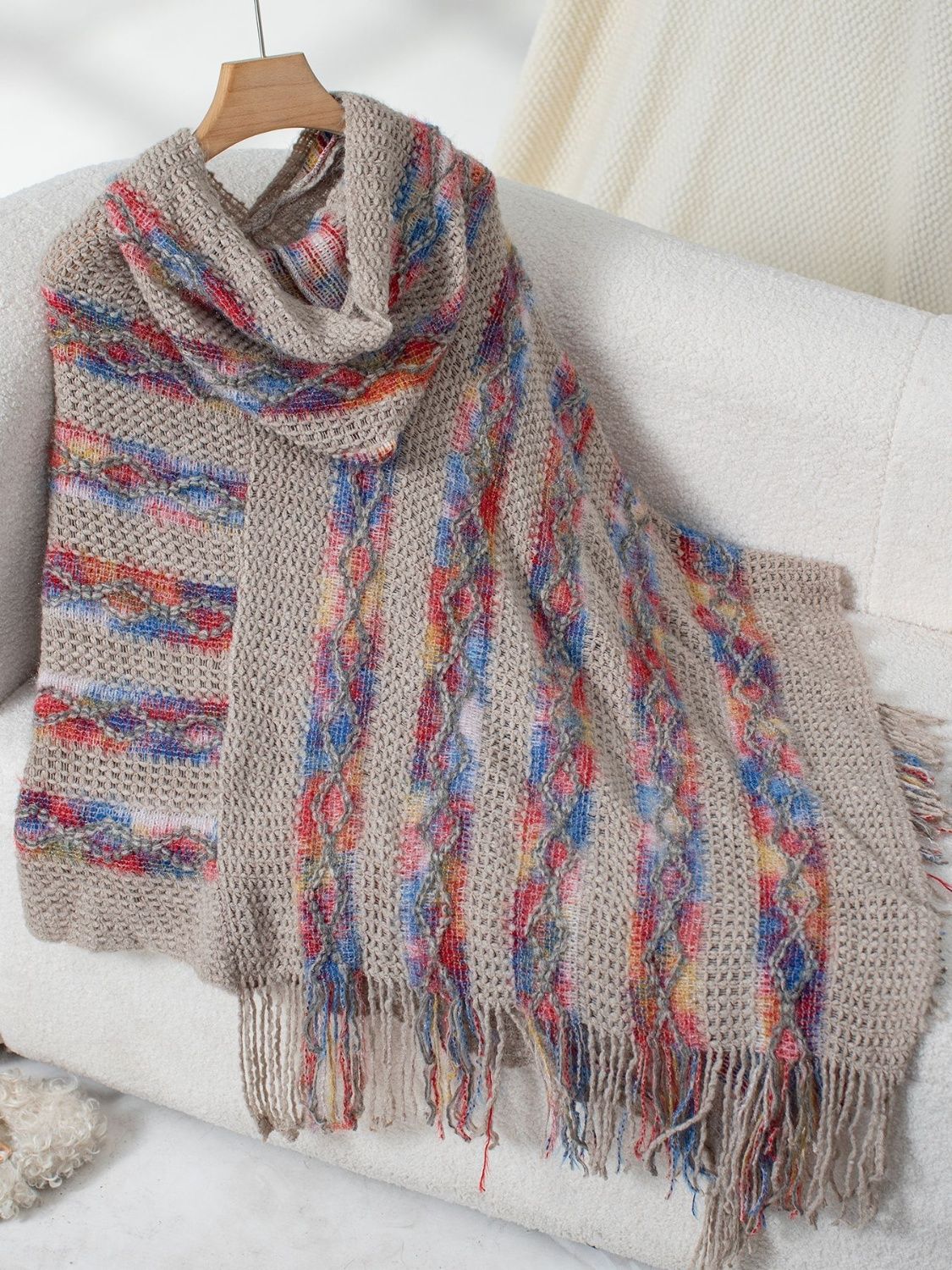 Textured knit hooded cape in beige with colorful striped and cable knit details and fringe.
