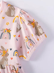 Pink cotton baby dress with playful bunny and rainbow print, puff sleeves, and ruffled hem.