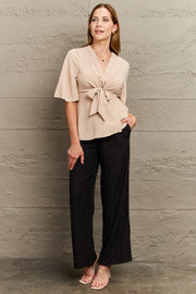Light apricot wrap-front blouse with tie waist and flutter sleeves, styled with black pants.