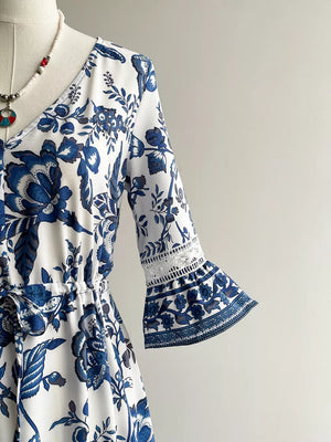 White and blue floral maxi dress with V-neck, drawstring waist, and lace-trimmed bell sleeves.