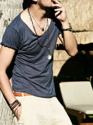 Men's dark blue cotton scoop neck tee with rolled sleeves, worn with tan pants and jewelry.