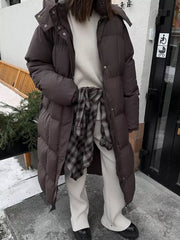 Dark brown padded trench coat with hood and tie-belt, worn open over a cream sweater and plaid shirt.