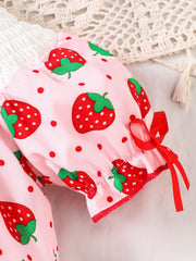 Baby pink one-piece with strawberry print, smocked detail, and red ribbon bows on sleeves.