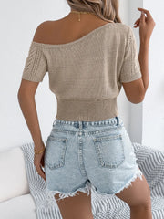 Tan open-knit crop top with asymmetric neckline and short sleeves, worn with denim shorts.
