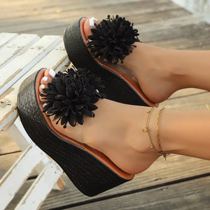 Black floral pom-pom wedge sandals with clear straps and a chunky platform sole.