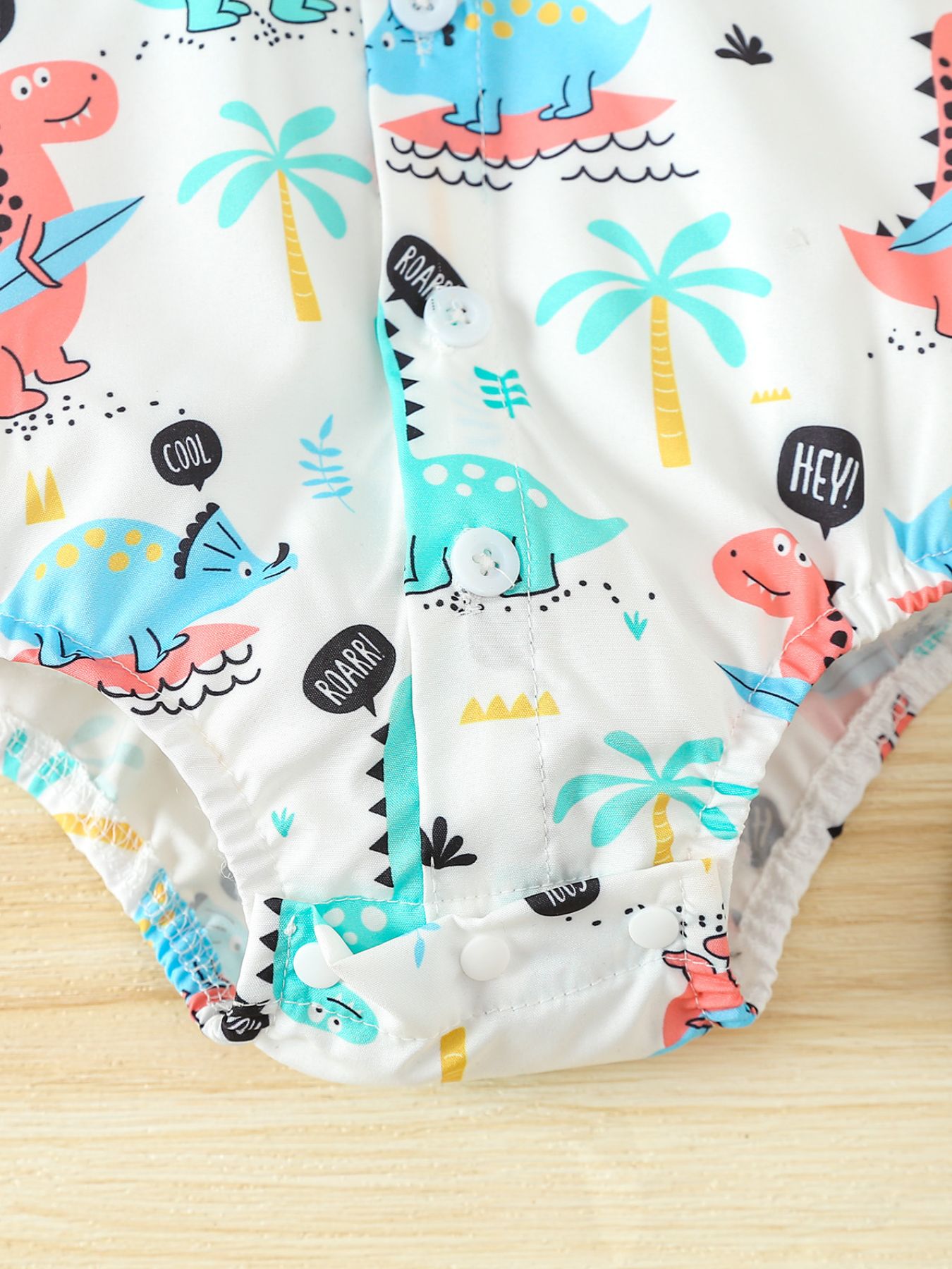 White cotton baby bodysuit with colorful dinosaur print, palm trees, and speech bubbles.