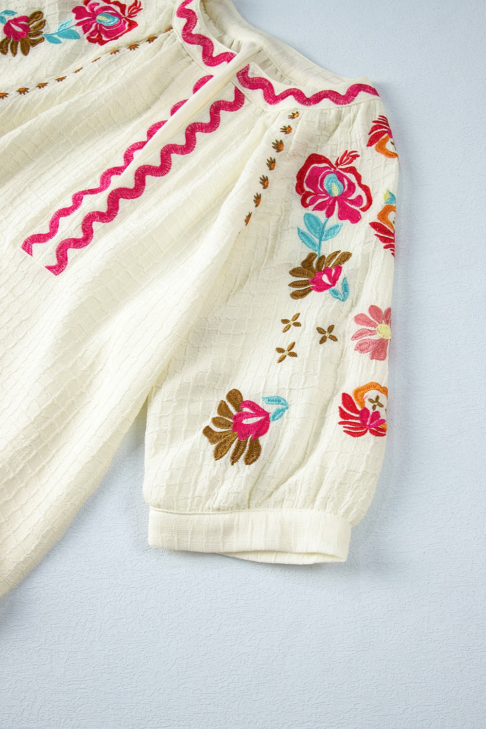 Beige textured blouse with embroidered floral sleeves and pink zigzag trim detail.