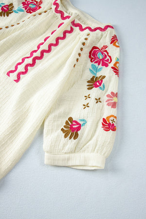 Beige textured blouse with embroidered floral sleeves and pink zigzag trim detail.