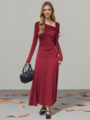 Burgundy long-sleeve maxi dress with asymmetrical neckline and tie detail.