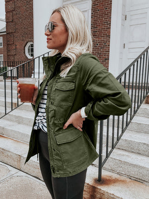 Army green utility field jacket with multiple pockets, zipper and button closure, worn casually.