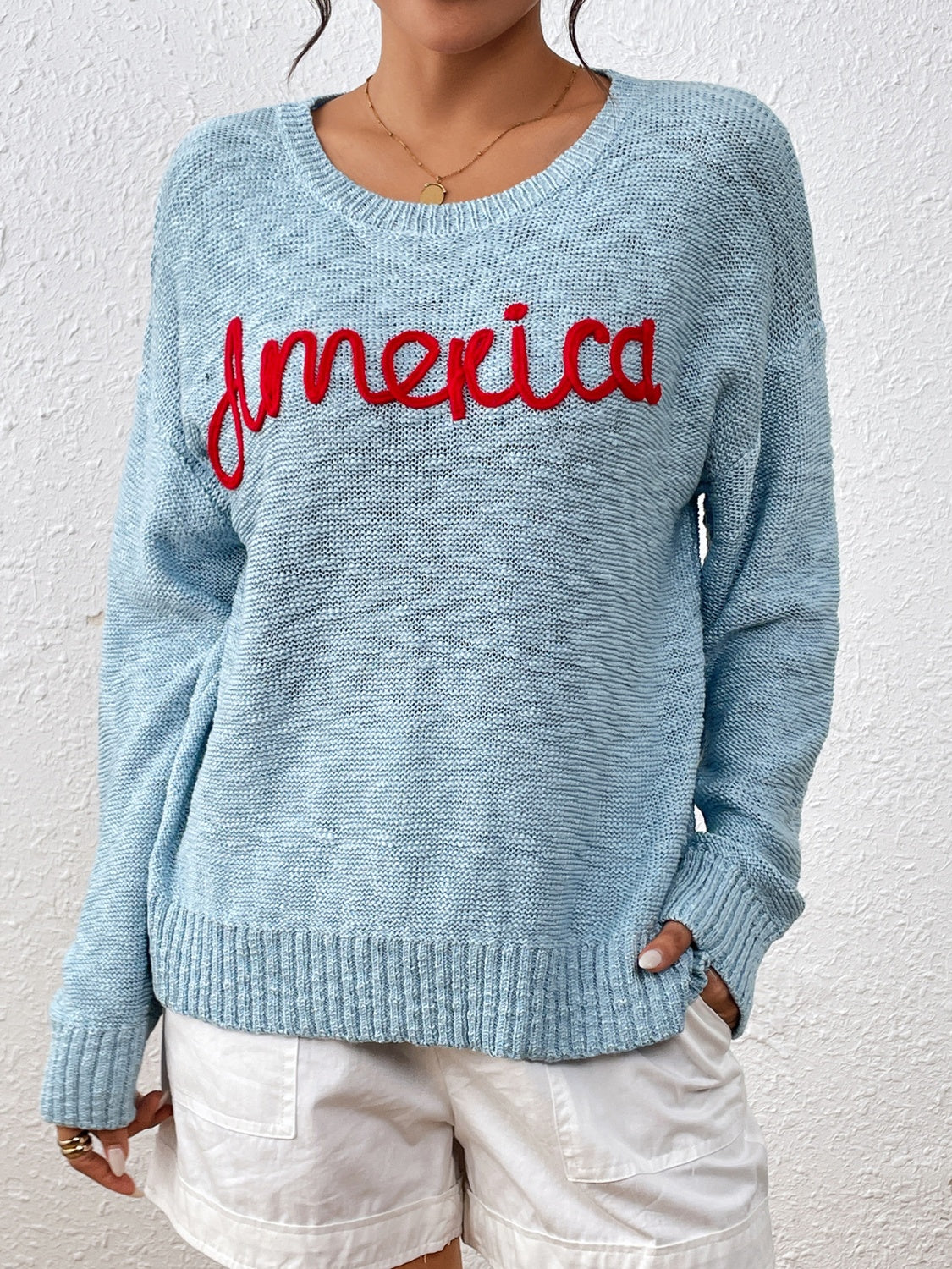 Pastel blue knit sweater with red embroidered "America" text, crew neck, long sleeves.