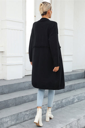 Black longline knit cardigan with fringe trim detail and pockets, worn open.