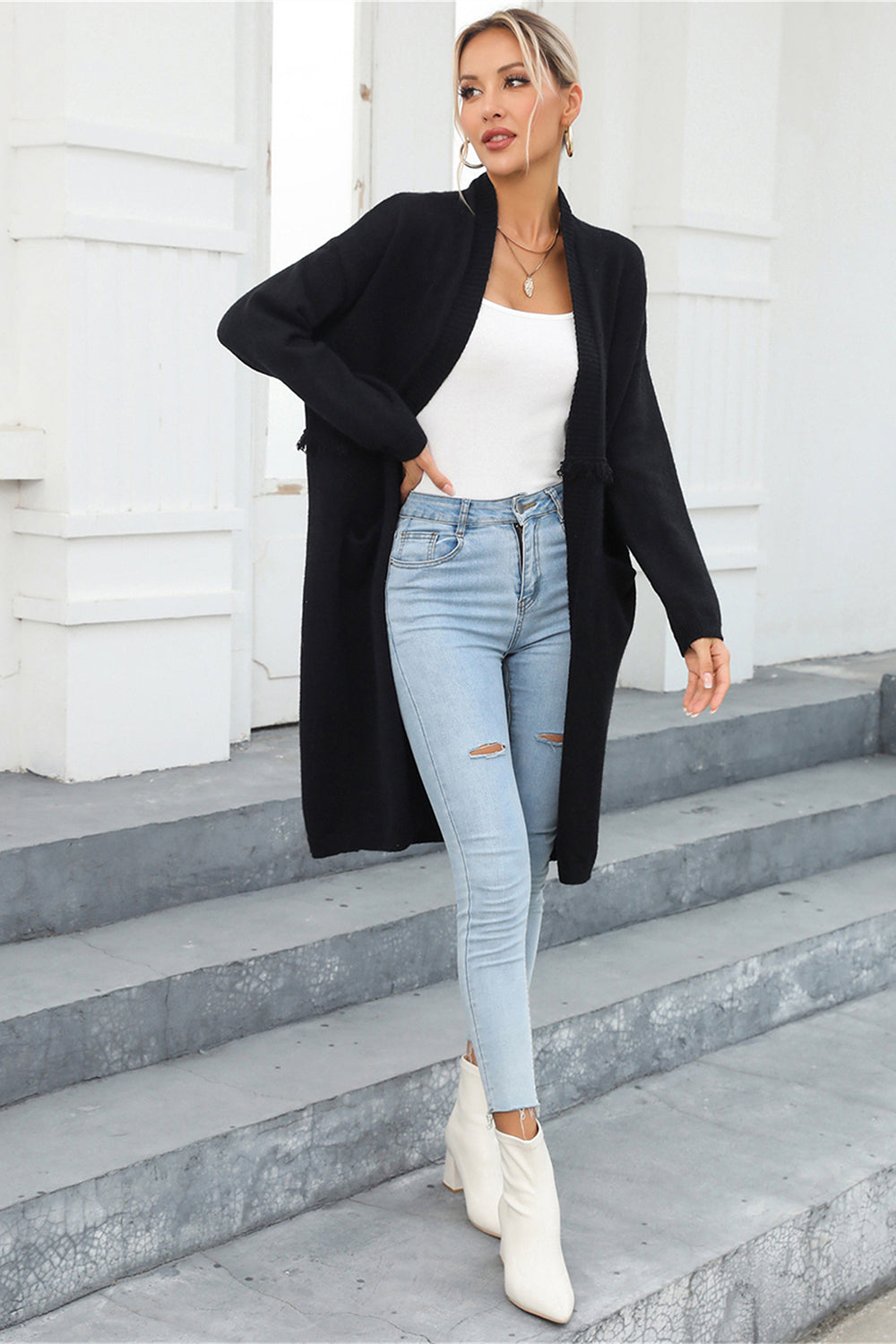 Black longline knit cardigan with fringe trim and pockets, worn open over a white tank top and ripped jeans.