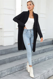 Black longline knit cardigan with fringe trim and pockets, worn open over a white tank top and ripped jeans.
