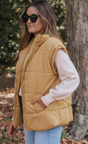 Tan padded sleeveless hooded vest with pockets, worn over a pink sweater.