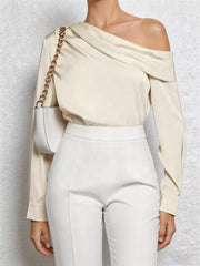 Chic cream one-shoulder draped blouse with a gold chain detail, styled with white pants.
