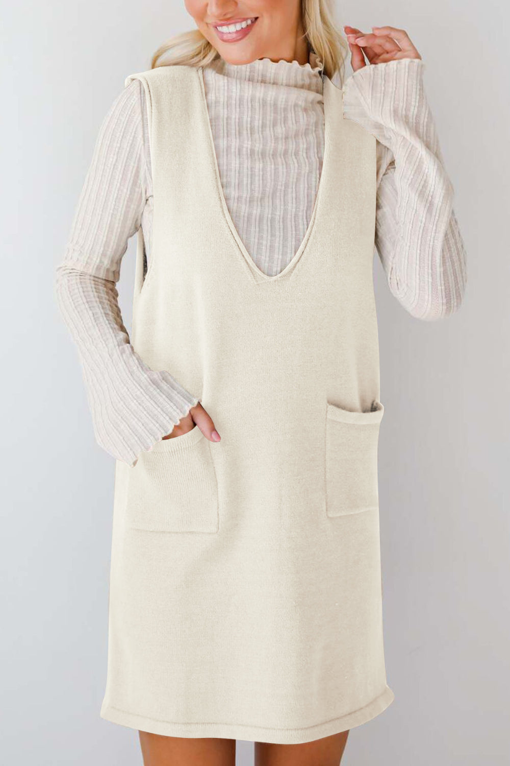 Cream sleeveless knit V-neck dress with two front pockets, layered over a ribbed turtleneck.