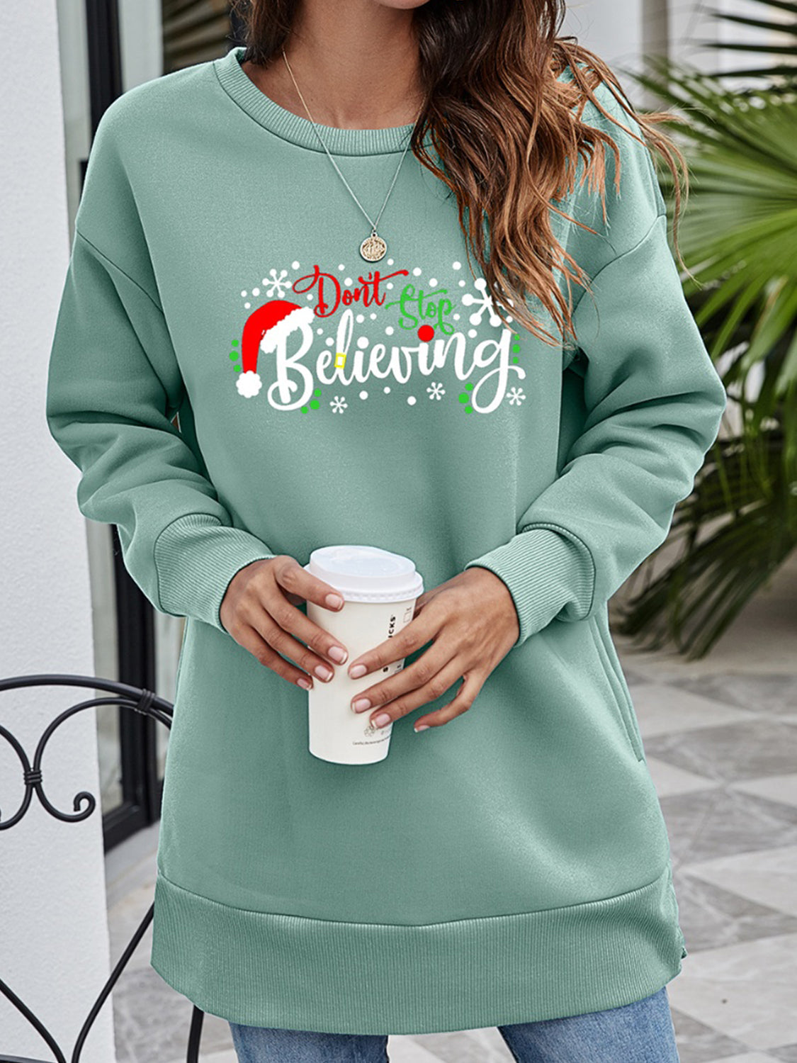 Sage green graphic tunic sweatshirt with "Don't Stop Believing" holiday print and Santa hat detail.