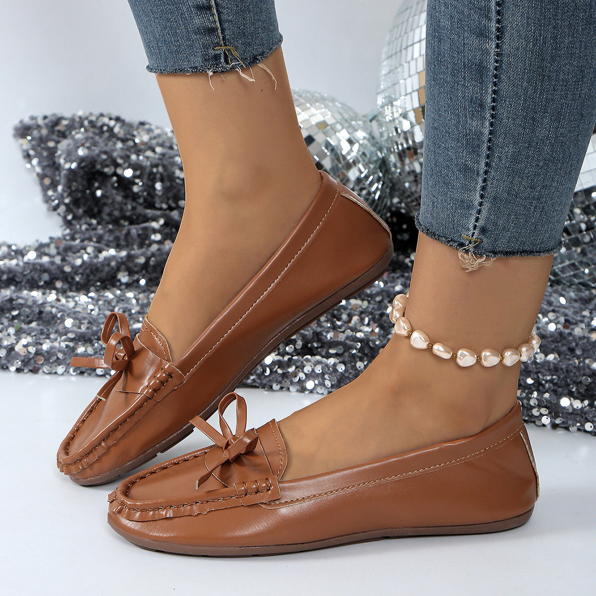Brown bow embellished flat slip-on loafers with decorative stitching and a pearl anklet.