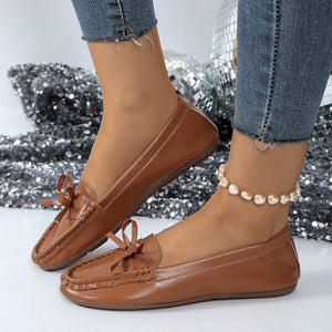 Brown bow embellished flat slip-on loafers with decorative stitching and a pearl anklet.