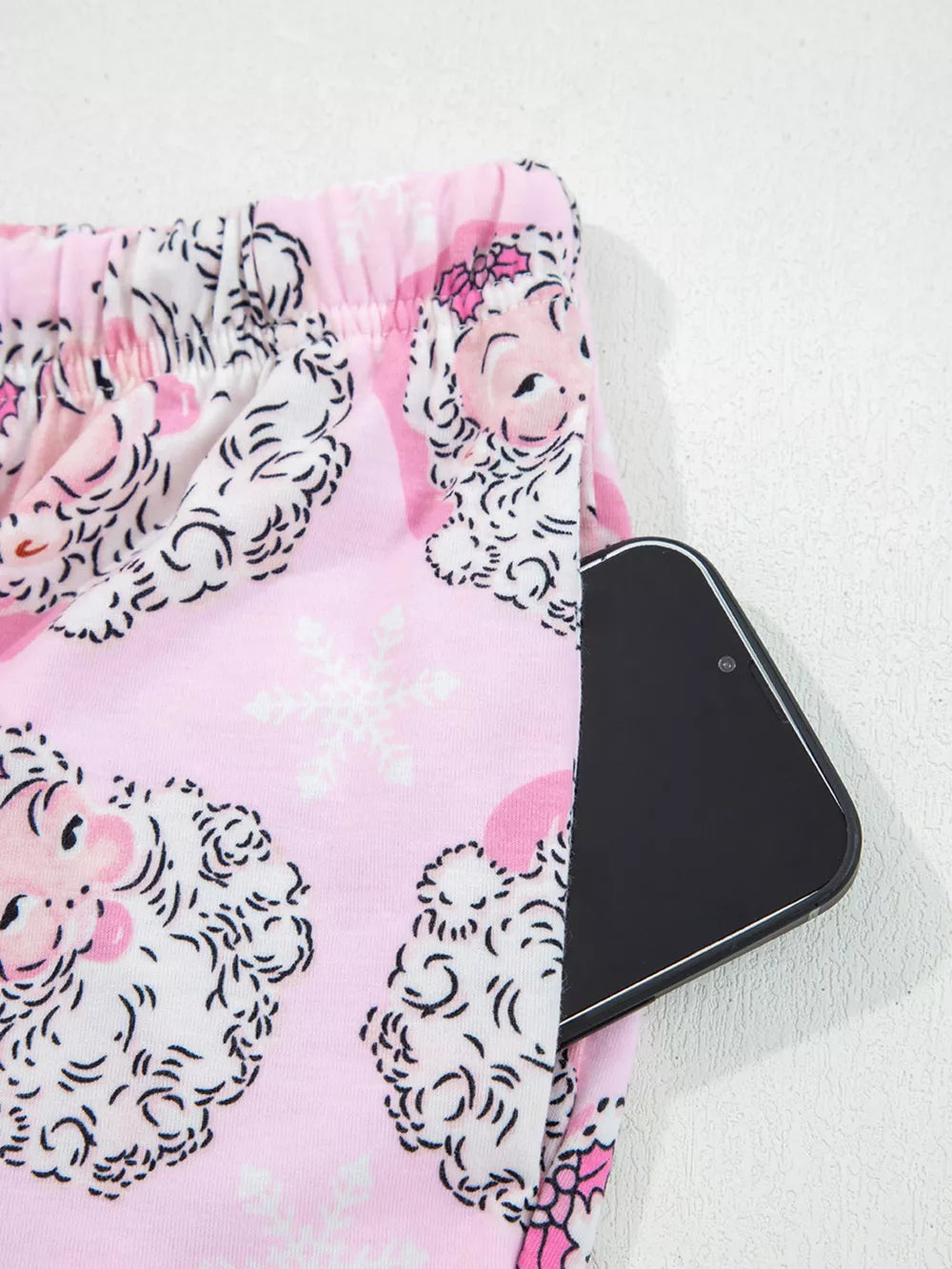 Pink pajama pants with Santa print and snowflake pattern, featuring a side pocket holding a smartphone.